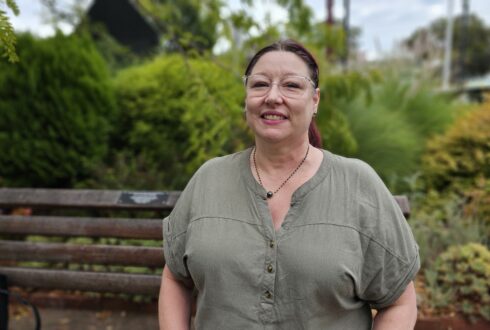 Rosy stands smiling in front of a park bench and some greenery and plants. She wears a short sleeve khaki blouse.