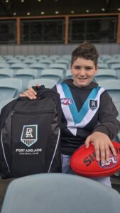 Lachlan sits smiling at the camera holding a football and wearing Port Adelaide Football Club colours