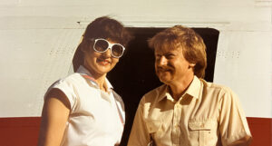older photo of woman in sunglasses standing next to man in front of plane