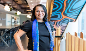 Woman with black hair and a blue scarf around her black t-shirt stands smiling in office setting