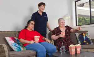 2 women sitting on couch watching tv with popcorn, as support worker stands behind them