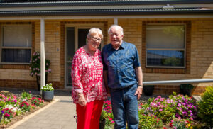 An elderly couple standing outside their retirement living unit, smiling.