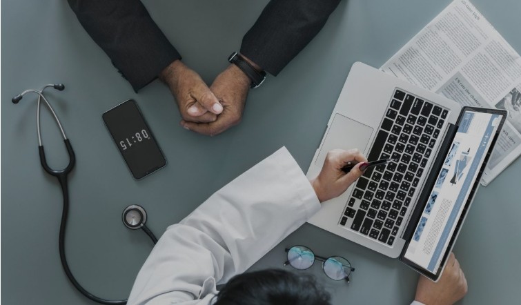 Image of doctor pointing to their laptop screen and the hands of the person listening can also be seen in the picture. 