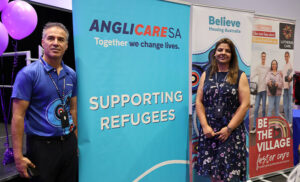 Man and woman stand beside a teal blue sign reading "Supporting refugees"