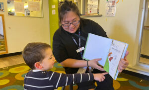 childhood educator in black tshirt reads a book to young boy pointing at page