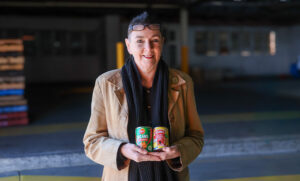 AnglicareSA volunteer Lisa smiling and holding tinned food donations at the warehouse where she sorts supplies for the community.