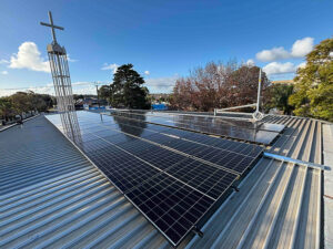 Solar panels installed on a church roof with a metal cross visible on the left side, photographed under a blue sky with surrounding trees in the background.