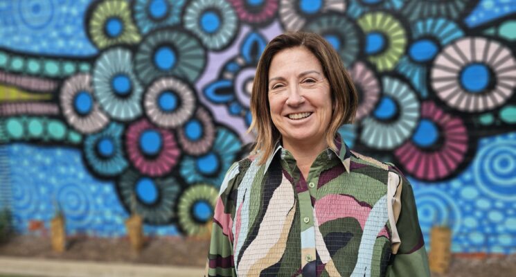 Catherine stands smiling in front of a blue coloured Indigenous mural.