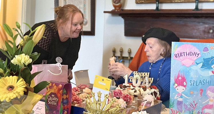 Leah, wearing a beret, and mid-blue jumper, celebrates her 104th birthday surrounded by flowers, gifts, cards and a cake.