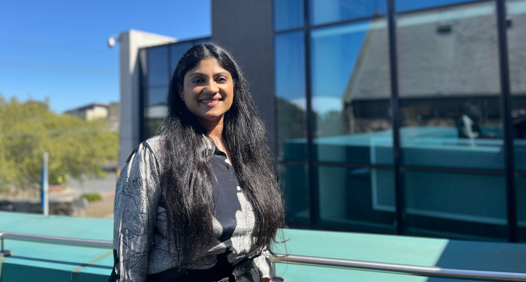 Prerna smiles at the camera. She is standing outisde on a balcony. She has long black hair and is wearing a black and grey flowery shirt.