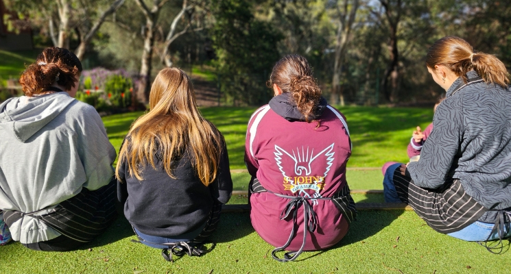 Four Students sit on a grassy oval wearing aprons with gum trees in the background.