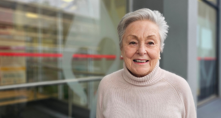 An older woman is smiling, wearing a dusty rose pink top and pearl earrings and standing in front of a glass office front.