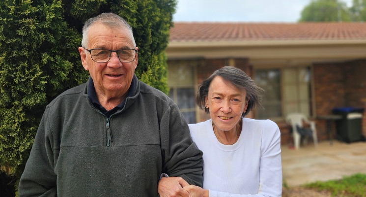 An older couple stands together in front of a home.