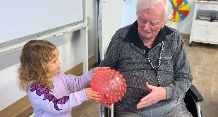 A small toddler dressed in a purple playsuit passes a red ball to an older man dressed in a grey jumper.