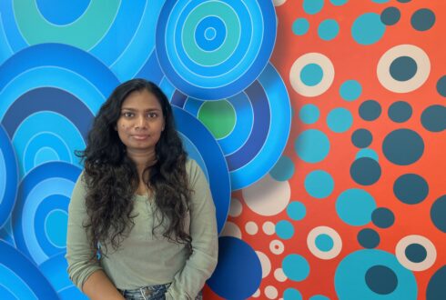 Ashini Pamudi, EA volunteer stands smiling in front of a brightly coloured mural. She wears a green shirt and has long dark hair.