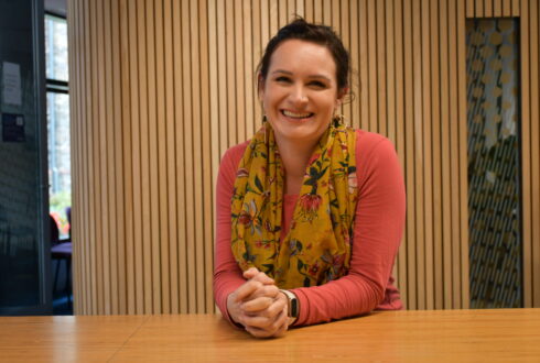 Jenna leans with her elbows on a table. She smiles and wears a pink long-sleeved top and yellow floral scarf.