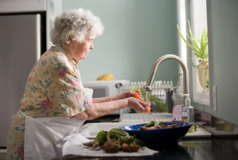 Older woman washing vegetables at kitchen sink.