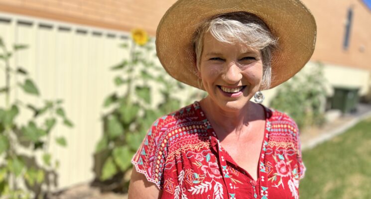 Sophie Thomson stands smiling in a garden wearing a straw hat and red blouse.