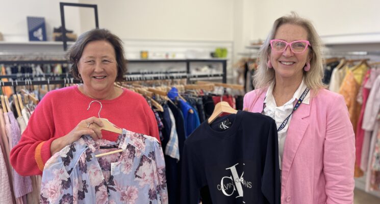 Mary-left-and-Maggie-right-Thread-together-volunteers stand smiling in front of racks of clothing.