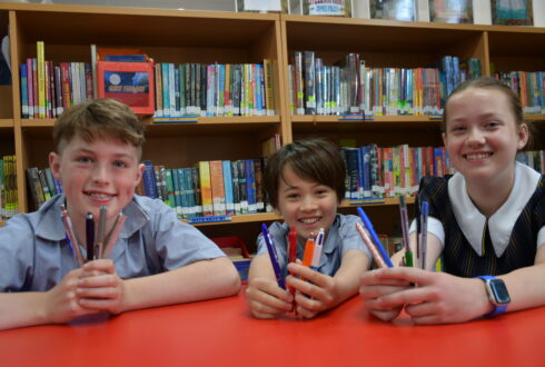 Pulteney Grammar students Max, Lily and Ashton sit in the library smiling and holding pens.