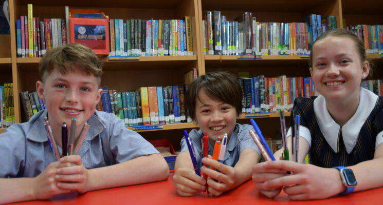 Pulteney Grammar students Max, Lily and Ashton sit in the library smiling and holding pens.