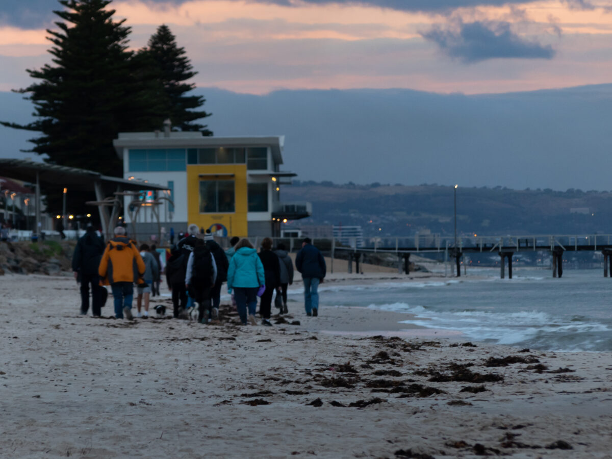 A group of people walk towards Henley Beach jetty as the sun rises in the background. 